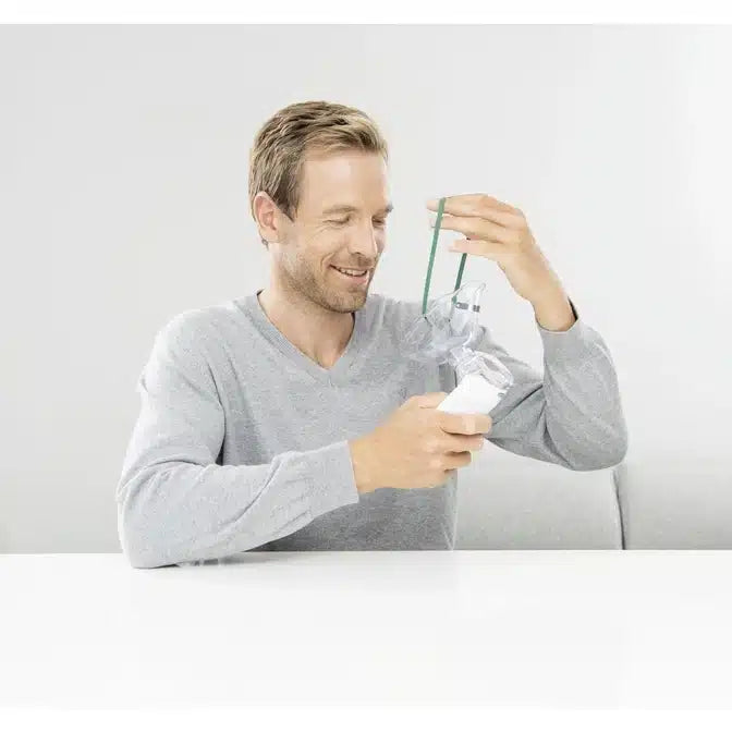 Ein Mann sitzt in einem hellgrauen Pullover an einem Tisch und benutzt einen Beurer Inhalator To Go IH 5 der Beurer GmbH. In seiner rechten Hand hält er die transparente Maske, die mit einem grünen Schlauch verbunden ist, während er mit der linken den Schlauch einstellt. Er lächelt leicht, seine Augen sind teilweise geschlossen. Der Hintergrund ist schlicht weiß.
