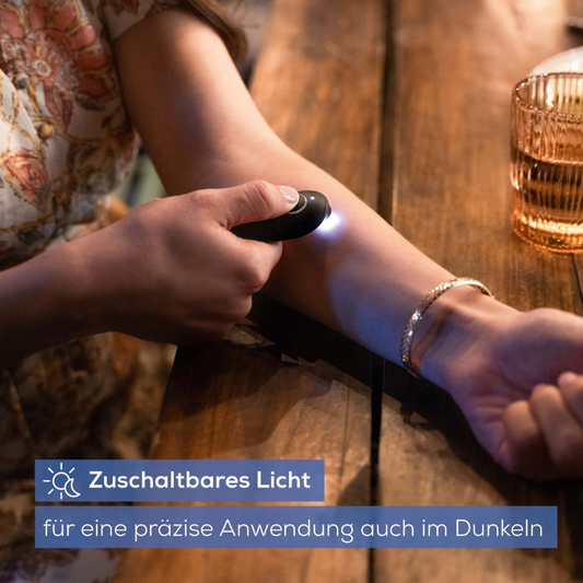 Eine Person hält einen schwarzen Beurer Insektenstichheiler BR 90 der Beurer GmbH in der Hand, eine kleine Lampe beleuchtet ihren Unterarm. Sie sitzt an einem Holztisch, im Hintergrund ist ein gemusterter Stoff mit Blumenmotiv zu sehen. Der Text auf Deutsch lautet „Zuschaltbares Licht für präzisen Einsatz auch im Dunkeln.“