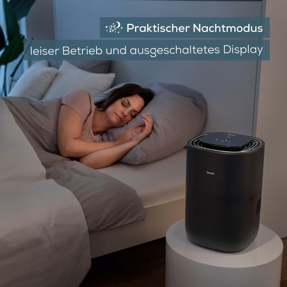 Eine Frau schläft im Bett, neben ihr steht ein Beurer LB 200 Luftbefeuchter mit Kaltverdunstungstechnologie auf dem Nachttisch. Das Bild ist mit einem Text in deutscher Sprache überlagert, der lautet: „Praktischer Nachtmodus, leiser Betrieb und ausgeschaltetes Display“. Der Raum ist schwach beleuchtet und der Luftbefeuchter ist aktiv.