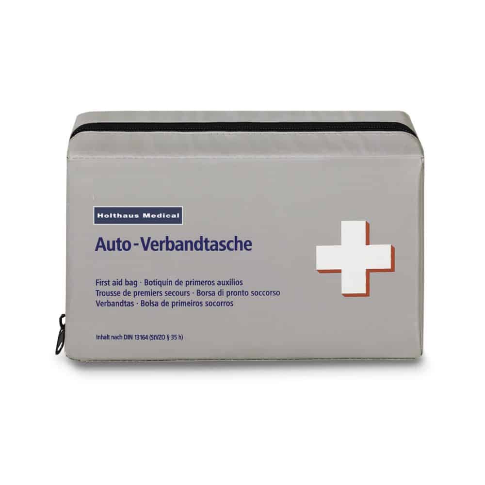 Eine rechteckige, graue Verbandtasche mit schwarzem Reißverschluss. Die Tasche trägt den Text „Holthaus Medical Klassik Verbandtasche“, „Auto-Verbands-Tasche“ und hat rechts ein weißes Kreuz mit rotem Rand. Oben ist sie mit der Marke „Holthaus Medical GmbH & Co. KG“ beschriftet, die der DIN 13164 entspricht.