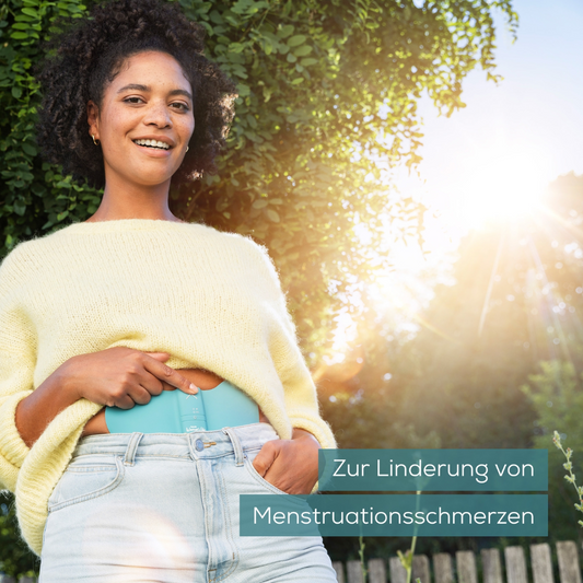 Eine Person in einem gelben Pullover und Jeans steht im Freien und hält ein Beurer EM 55 Menstrual Relax+ Gel-Pad der Beurer GmbH in der Hand. Im Hintergrund ist Grün zu sehen, durch das Sonnenlicht fällt. Der Text lautet „Zur Linderung von Menstruationsschmerzen“.