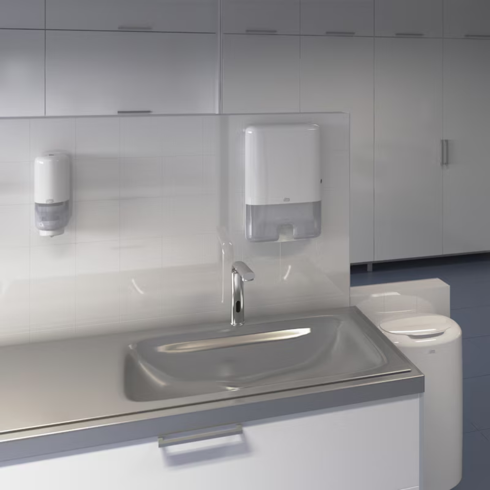 Ein minimalistischer Waschtischbereich im Badezimmer verfügt über ein Edelstahlwaschbecken mit einem modernen Wasserhahn. Über dem Waschbecken sind ein Seifenspender und ein Papierhandtuchspender an der Wand montiert, die ein Elevation-Design zeigen. Der Hintergrund zeigt weiße Schränke und einen TORK Tork 563000 Abfallbehälter Elevation 50 Liter B1 | Packung (1 Stück) zur Abfallentsorgung.