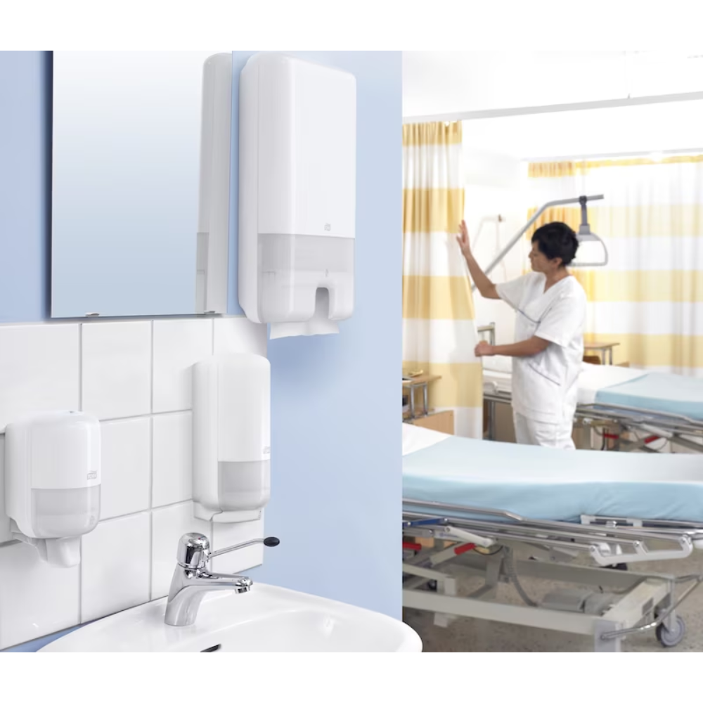 Ein Krankenhauszimmer mit einer blau-weißen Wand, an der Seifenspender, Tork Xpress® 552000 Spender für Multifold Handtücher H2 | Packung (1 Stück) von TORK und ein Waschbecken hängen. Im Hintergrund passt ein Mitarbeiter des Gesundheitswesens in weißer Uniform Geräte neben einem Bett mit blauen Laken und einem gelb-weiß gestreiften Sichtschutzvorhang an.